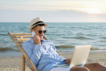 Happy man with laptop talking by mobile phone on beach. Business trip