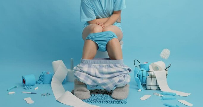 Unrecognizable woman suffers from abdominal pain sits on toilet bowl in lavatory takes medicines touches belly has diarrhea or constipation wears casual t shirt shorts blue panties and slippers.