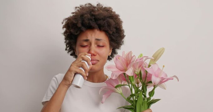 Unhappy dark skinned woman sneezes and has runny nose uses nasal spray reacts to allergen holds lilies dressed casually isolated over white background. Experiencing allergy. Seasonal allergic rhiniris