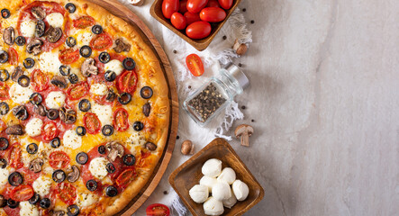 On a white background pizza on a wooden plate. Top it is decorated with tomatoes, olives and mozzarella cheese. Tomatoes and mushrooms in wooden bowls on the table. View from above.
