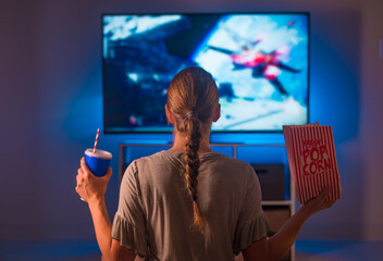 A young blonde woman in pajamas sits in front of a TV screen in the bedroom and watches her favorite movie. She is holding a pack of popcorn and a drink with a straw in a plastic glass. - 447986650
