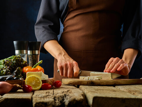 Healthy eating. Fitness. Sport. The chef prepares a delicious low-calorie Greek salad dish. He is slicing feta cheese on a cutting board. Other healthy and tasty ingredients are on the table.