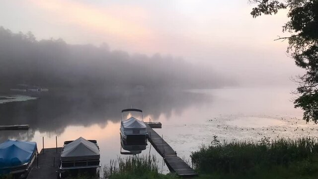 An early morning lakeside time lapse shot of pond fog wafting and dissipating over lily pads, a dock, and covered pontoon boats at Buskey Bay, Iron River, Wisconsin.