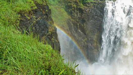 Water streams fall into the gorge. A rainbow formed between the stony slopes. Green grass in the...
