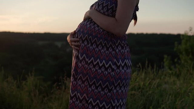 happy pregnant woman touching her tummy on top of mountain in tall grass before sunset. caring mother strokes large belly tummy with hands. pregnancy, motherhood, people, expectation, summer concept