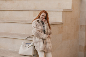 Fashion portrait of young stylish lady posing near stairs outdoors. Charming red-haired girl with beige plaid coat and trendy bag, smiling, touching chin and looking into camera