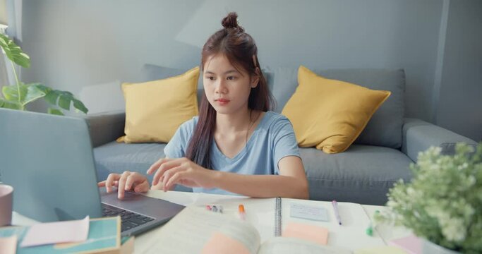 Young Asia girl teenager with casual use laptop computer learn online write lecture notebook for final test in living room at house. Isolate education online e-learning coronavirus pandemic concept.