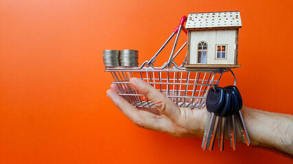 Buying and selling real estate. Rent an apartment. Capital investments in residential buildings. Coins and keys to a wooden house in a supermarket basket held by a human hand. Orange background.