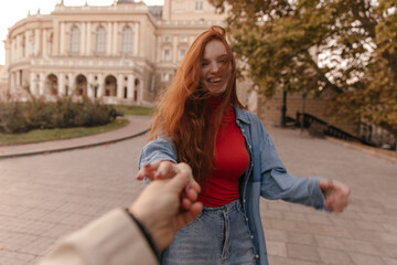 Street photo of young lady standing against city landmark background. Attractive long-haired ginger in red top and denim shirt, posing outdoors and smiling