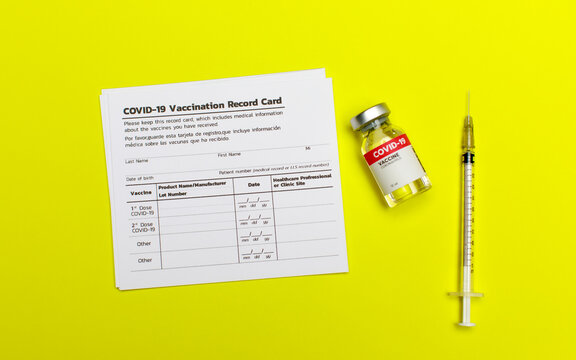 Covid-19 vaccination record card including info of patient, dose, product lot, injection date, clinic, healthcare professional beside vaccine glass and medical syringe.