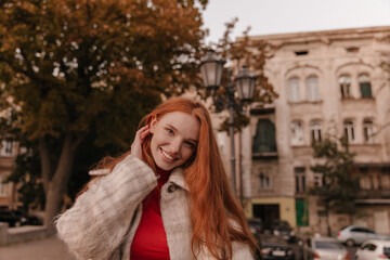Naklejka na ściany i meble Lifestyle portrait of young ginger posing in city center. Pretty long-haired girl with stylish outfit, adjusting hairstyle and smiling outdoors