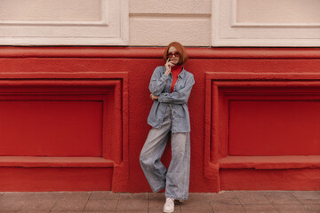Street fashion portrait of young lady standing against bright building background. Trendy red-haired girl, wearing denim outfit and white sneakers, adjusting sunglasses and posing outdoors