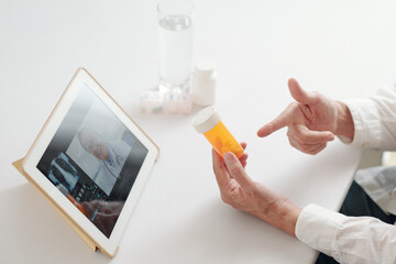 Close-up of doctor sitting at the table and pointing at bottle with medicine while having online meeting with his colleague on digital tablet