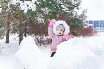 little girl in a warm coat and a hood plays snowballs in a snow fortress