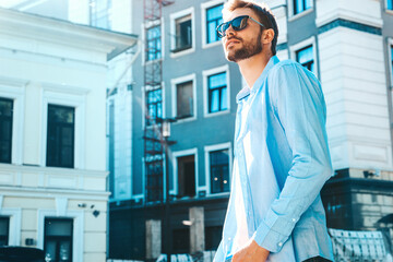 Portrait of handsome smiling stylish hipster lambersexual model.Modern man dressed in blue shirt. Fashion male posing near skyscraper on the street background. Outdoors at sunset