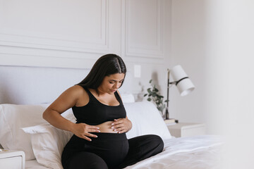 Pregnant Carribbean Indigenous woman practicing self care and putting lotion on baby bump in bed