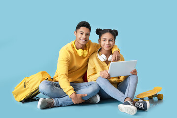 African-American brother and sister with laptop, skateboard and schoolbag on color background