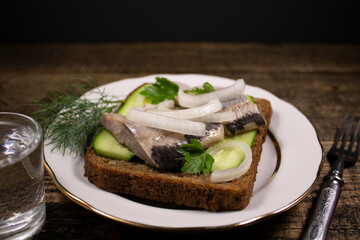 A sandwich of rye bread with salted herring.A delicious snack on a white plate,a fork and a glass of vodka.