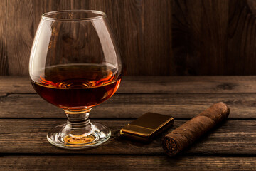 Glass of brandy and cuban cigar with golden lighter on an old wooden table. Focus on the cuban cigar