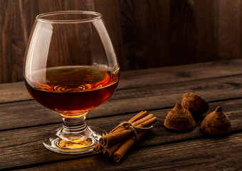 Glass of brandy and a couple of chocolate truffles with cinnamon sticks tied with jute rope on an old wooden table. Focus on the glass of brandy