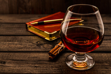 Glass of brandy and leather notebook with golden pen and cinnamon sticks tied with jute rope on an...
