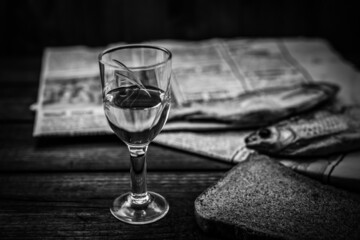 Glass of vodka with a stockfish and newspaper with piece of the black bread on an old wooden table. Angle view
