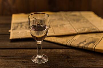 Glass of vodka and newspaper on an old wooden table. Angle view, focus on the glass of vodka