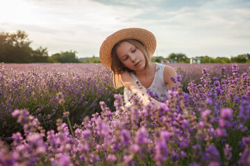 Teenage girl enjoys the scenery of lavender field. Dreamy teenager in straw hat sitting among purple flowers. Calm landscape, escape to beauty of nature, summer lifestyle