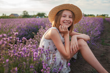 Teenage girl enjoys the scenery of lavender field. Dreamy teenager in straw hat sitting among purple flowers. Calm landscape, escape to beauty of nature, summer lifestyle