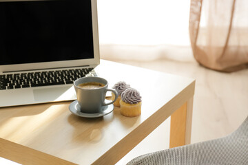 Table with cup of coffee, cupcakes and laptop in room