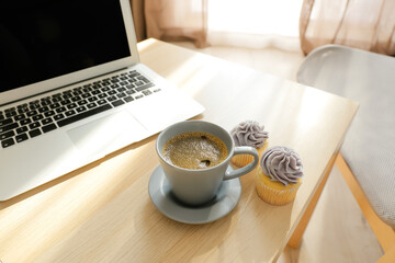 Table with cup of coffee, cupcakes and laptop in room