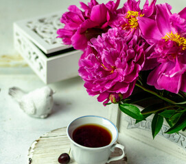 Shabby chic tea. A cup of tea on a white wooden tray with a cherry nearby. Peonies are in a white box. Blurred background
