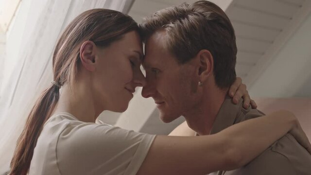 Tilting up of young Caucasian woman and man wearing loungewear, sitting at home in front of each other, holding in arms, touching foreheads