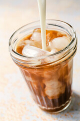 Pouring of milk into glass of hot coffee with ice on light background
