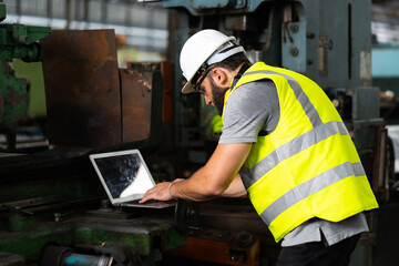 Young caucasian engineering man in Hard Hat Wearing Safety Jacket working on laptop computer in Heavy Industry Manufacturing Facility.
