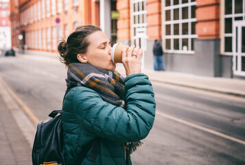 Young woman drinking coffee from a paper cup while standing on the street in the city on an autumn day