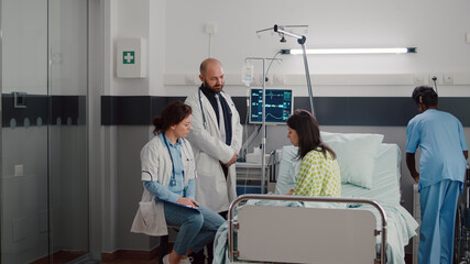 Obraz premium Specialist practitioner doctors monitoring sick man explaining illness treatment while working in hospital ward. Patient sitting in bed discussing symptom disease during recovery appointment