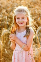 A beautiful little blonde with long hair in a pink dress stands on a wheat field in the rays of the setting sun