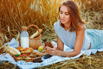 A young woman on a picnic in the countryside. A girl is lying on a blanket with snacks, bread, milk and fruit in a wheat field at sunset. © Elena 