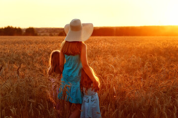 A daughter holds her mother's hand, stands on a golden wheat field at sunset. A young woman and two girls in dresses are standing among the rye. Agro walk in the countryside