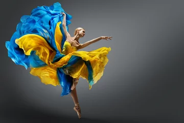Fotobehang Mooie vrouw balletdanser springen in de lucht in kleurrijke fladderende jurk. Sierlijke ballerina danst in geelblauwe jurk op grijze studioachtergrond © inarik