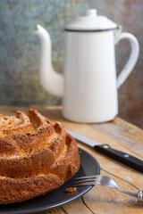 Close-up of yogurt sponge cake on dark plate, with wooden fork, knife and old white coffee pot, on wooden table, selective focus, vertical