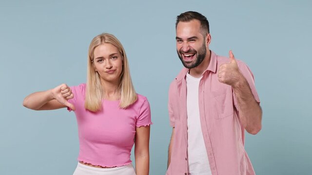 Confused smiling young couple two friends family bearded man woman in pink clothes together show thumb up down no like dislike gesture isolated pastel plain light blue color background studio portrait
