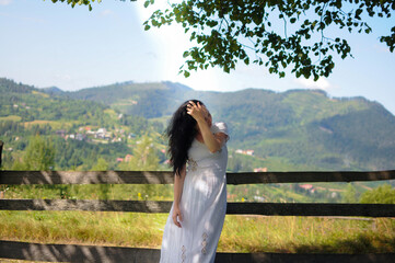 Young woman in white dress is standing near a fence on mountain meadow.