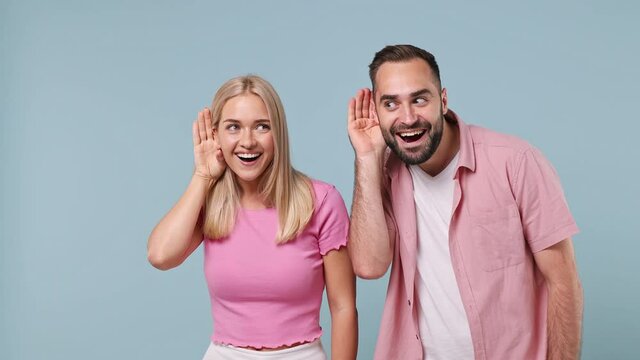 Young couple two friends family man woman in pink clothes look aside hold hand near ear try to hear you overhear listen intently isolated on pastel plain light blue color background studio portrait