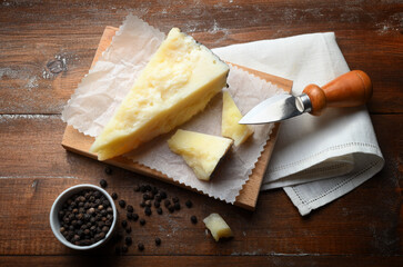 Pieces of pecorino cheese on cutting board with white napkin, hard cheese knife and black peppercorns. Rustic wooden background, high angle view.