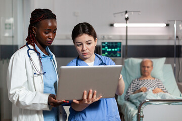 African american doctor and nurse using laptop in hospital room for sick senior patient diagnosis...