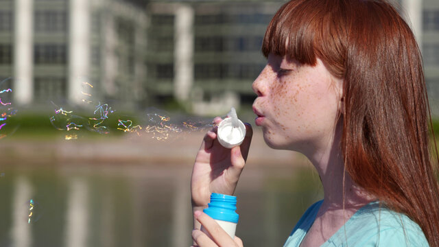 Side view of teenage redhead girl blowing soap bubbles outdoors.