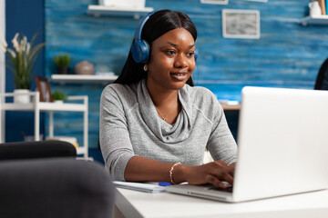 African american student wearing headphones browsing information composing blog article using laptop computer. Black woman typing education email working at social media webinar in living room