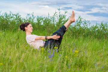 Beautiful young woman practices yoga outdoors. Sits on the ground with legs raised and holds a corner.